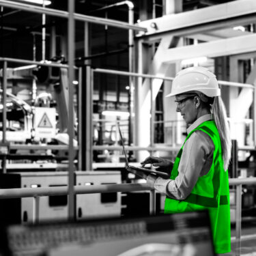 Car Factory: Female Automotive Engineer Wearing Hard Hat, Standing, Using Laptop. Monitoring, Control, Equipment Production. Automated Robot Arm Assembly Line Manufacturing Electric Vehicles.